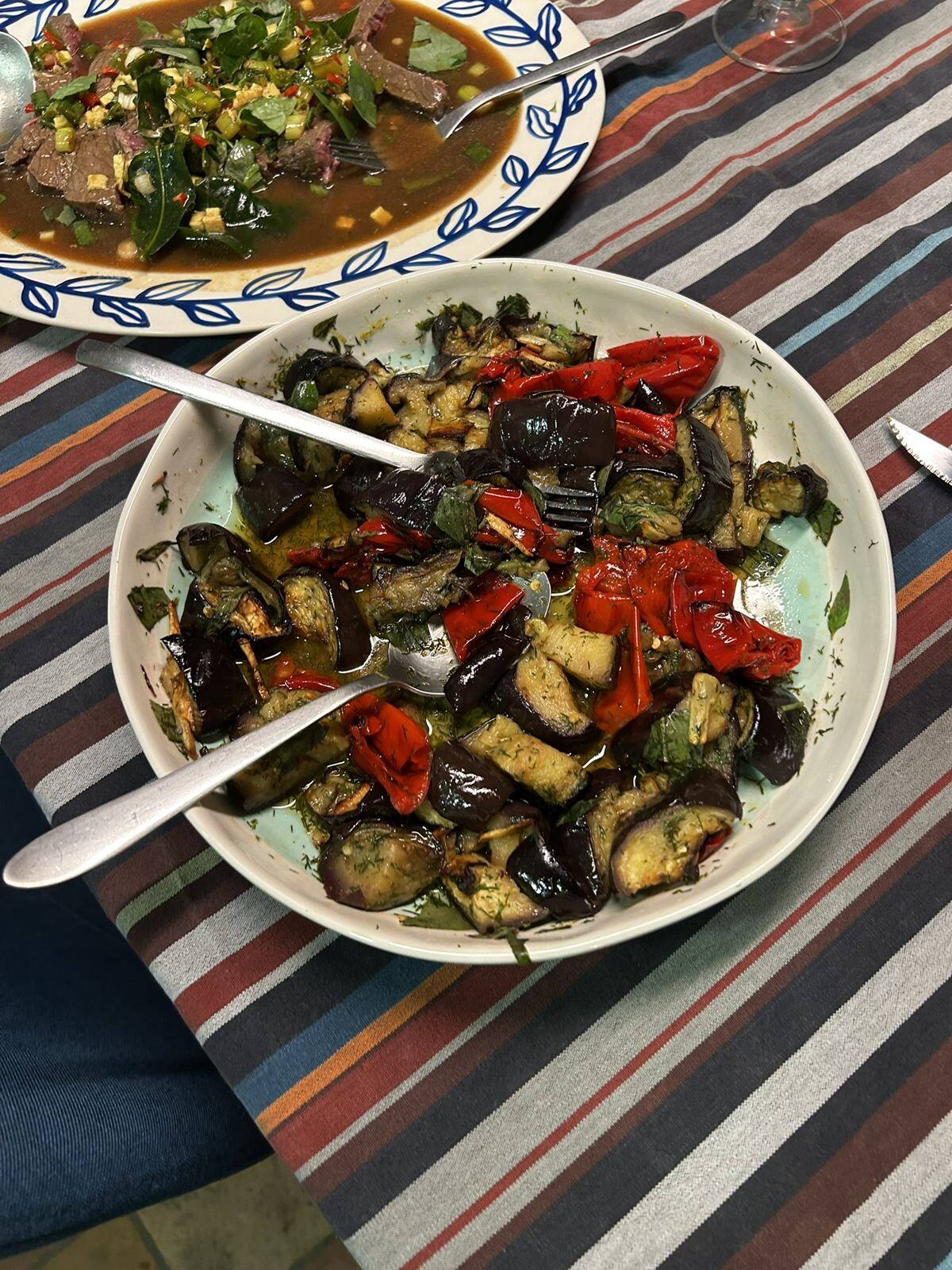 Sur une nappe à rayures, un plat d'aubergines rôties et de poivrons rouges à l'aneth, et dans le coin une assiette de bœuf aux herbes, au piment et au citron vert.