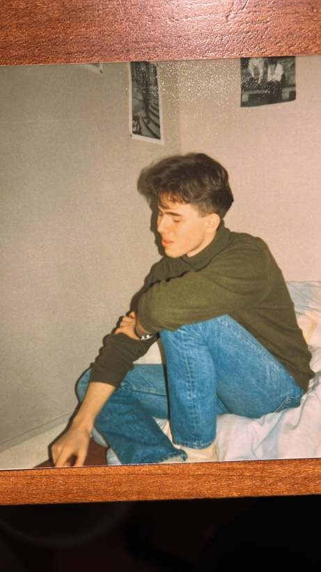 Un jeune homme en col roulé vert, assis sur un lit, les yeux baissés.