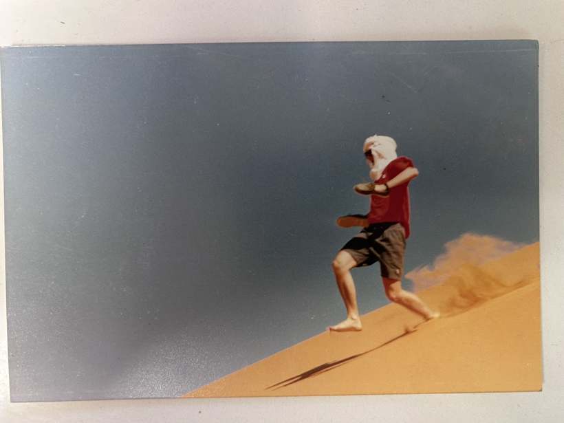 Un adolescent en T-shirt rouge, le visage couvert d’un chèche, dévale une dune.